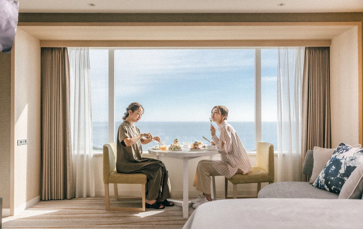 Two people enjoying room service in a room overlooking the sea.