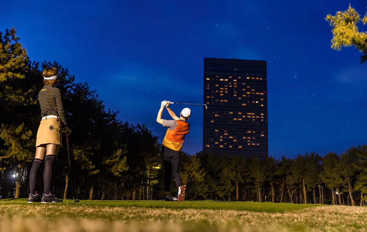 Two people hitting shots on a golf course at night.