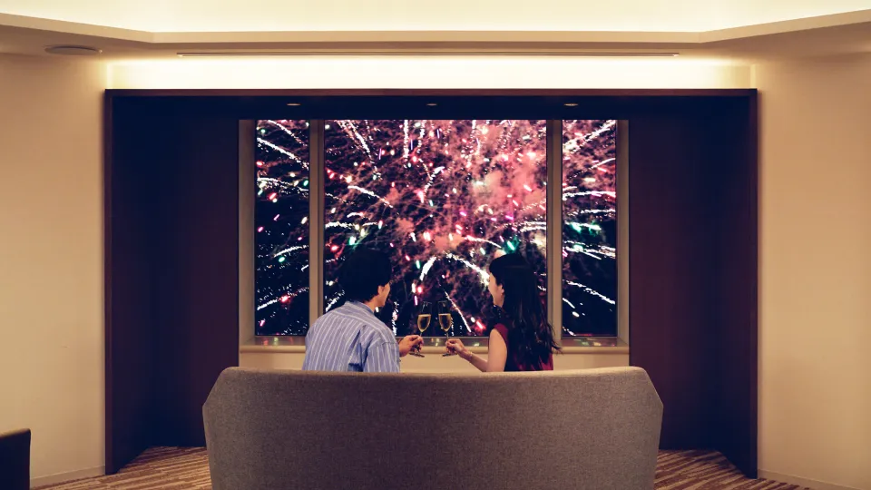 A man and a woman watch fireworks from the window of their hotel room.